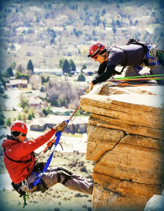 Two rock climbers wearing helmets on a ledge one is assisting another with ropes